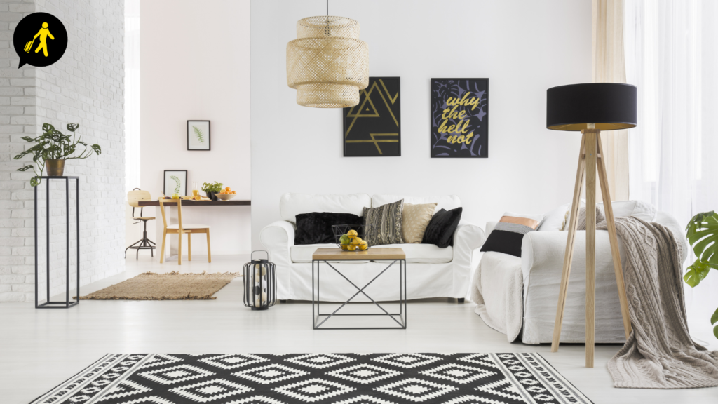 Photo shows an open plan living space with white walls and floor. It includes a white sofa, with black cushions, a minimalist coffee table, a small office desk, decorative lamps and a black and white patterned rug. This is an example of a servied accommodation.
